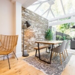 A general interior view of a dining room within an extension with glass conservatory roof, bare brick wall, oak dining table with metal legs, dining chairs, bench, rattan accent wing chair, industrial style floor lamp, oak floor looking into the garden through open bi-fold patio doors within a home