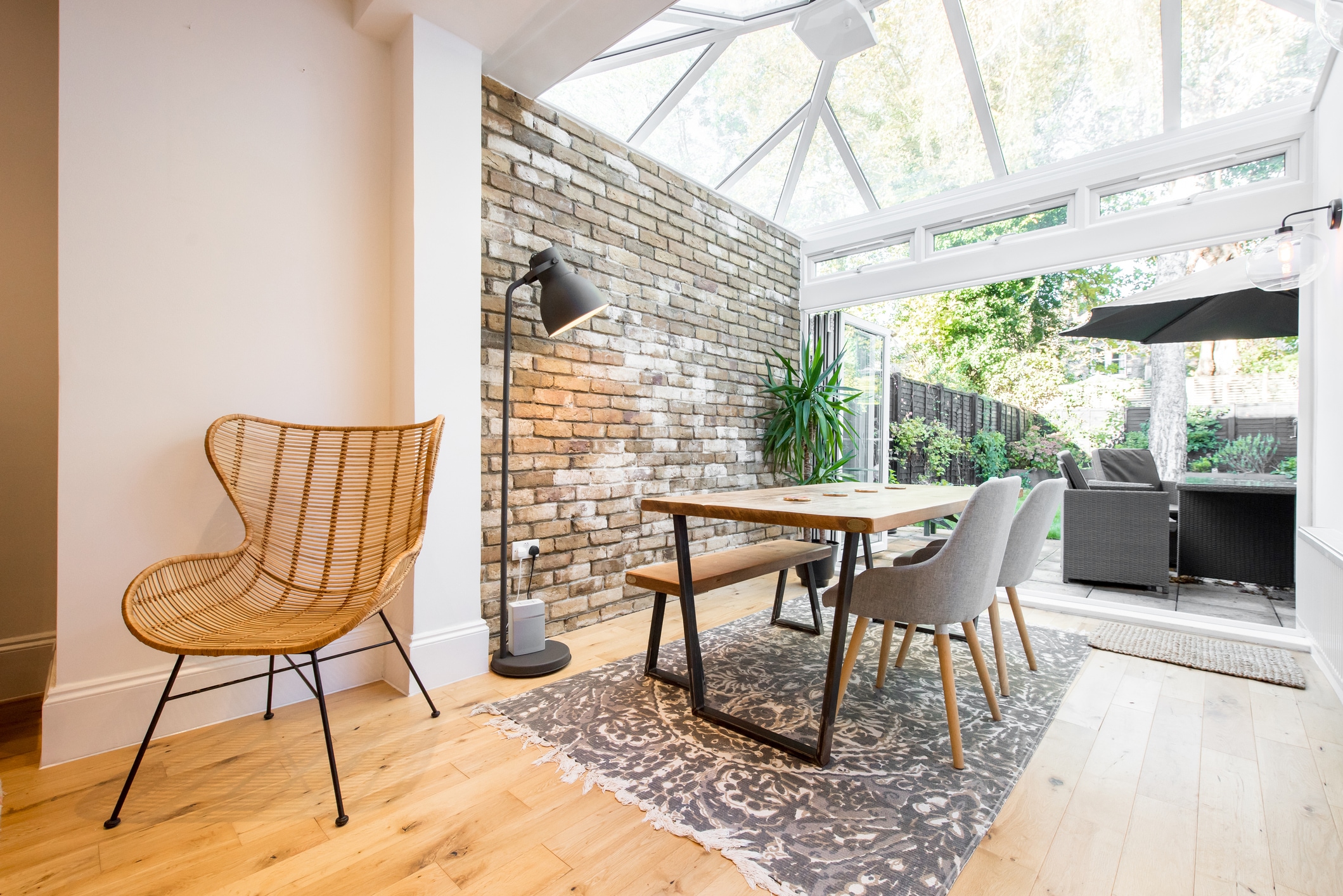 A general interior view of a dining room within an extension with glass conservatory roof, bare brick wall, oak dining table with metal legs, dining chairs, bench, rattan accent wing chair, industrial style floor lamp, oak floor looking into the garden through open bi-fold patio doors within a home