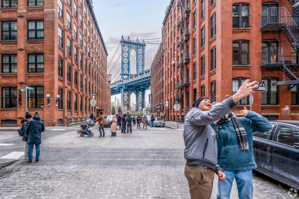 Two people take a selfie in the Vinegar Hill neighborhood of Brooklyn with the DUMBO Manhattan Bridge in the background.